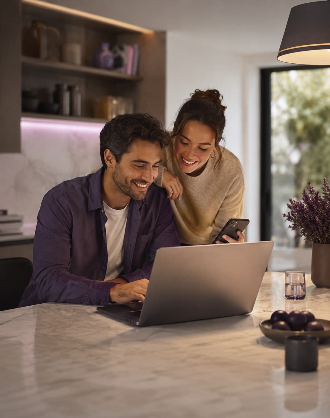 A couple smiling together at a kitchen counter, looking at a laptop and phone.